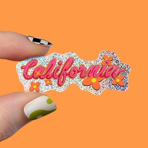 May include: A holographic sticker with the word "California" in pink script, adorned with orange flowers. The sticker has a glittery, iridescent finish and is set against an orange background.