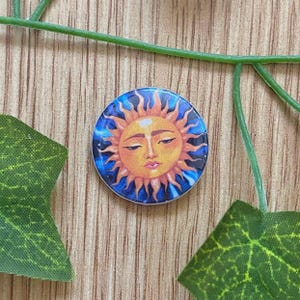 May include: A round, colorful button featuring a sun design. The sun has a yellow face with closed eyes, surrounded by orange rays against a blue background. The button is a decorative accessory.