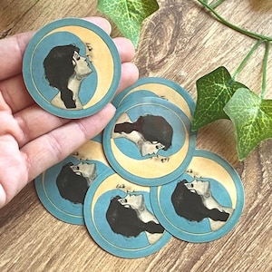 May include: A collection of round stickers featuring a woman's profile against a crescent moon. The stickers have a blue border and a cream-colored moon. The woman's hair is black, and the background is a light blue.