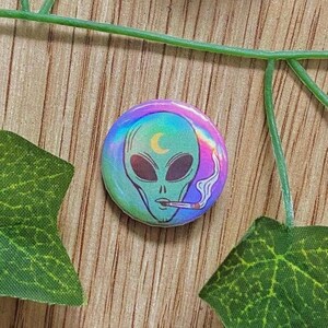 Könnte beinhalten: Ein runder Button mit einem grünen Alien, der eine Zigarette raucht, vor einem holografischen Regenbogenhintergrund. Der Alien hat große schwarze Augen und einen Mond. Rauch steigt von der Zigarette auf.