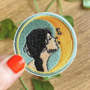 Könnte beinhalten: Gestickter Aufnäher mit dem Profil einer Person mit schwarzen Haaren und Schnurrbart, vor einem gelben und türkisfarbenen Hintergrund. Der Aufnäher hat eine kreisförmige Form mit einem gestickten Rand. Das Design beinhaltet Noten.