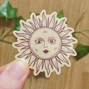 Könnte beinhalten: Ein beiger Aufkleber mit einem Sonnendesign mit Gesicht, Strahlen und holografischem Effekt. Das Gesicht der Sonne hat Augen, eine Nase und einen roten Mund. Der Aufkleber wird von einer Hand gehalten.