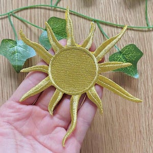 Könnte beinhalten: Gestickter goldener Sonnenaufnäher mit strahlenförmigen Strahlen. Die Sonne hat eine kreisförmige Mitte und spitze Strahlen, die sich nach außen erstrecken. Der Aufnäher besteht aus einem strukturierten Stoff und ist wahrscheinlich zum Aufnähen oder Aufbügeln auf Kleidung oder Accessoires bestimmt.