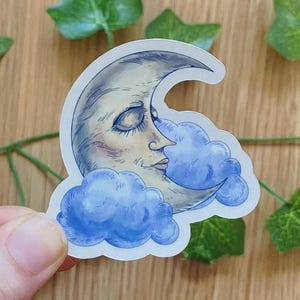 Può includere: Un adesivo raffigurante una luna crescente con un volto sereno, dipinto in tonalità grigie e crema. La luna è circondata da soffici nuvole blu. L'adesivo ha un bordo bianco ed è probabilmente decorativo.