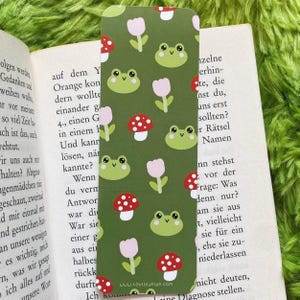 Könnte beinhalten: Ein grüner Lesezeichen mit einem Muster aus Cartoon-Fröschen, Pilzen und rosa Tulpen. Das Lesezeichen ist rechteckig und befindet sich in einem offenen Buch. Der Text "WWW.LOUISEKIND.COM" ist unten sichtbar.