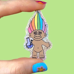 Könnte beinhalten: Glitzernder Aufkleber mit einem Zeichentrick-Troll mit Regenbogenhaar, der eine blaue Bong hält. Der Troll hat braune Haut und lächelt. Der Aufkleber wird zwischen Fingern vor einem grünen Hintergrund gehalten.