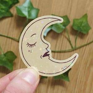 Puede incluir: Una pegatina con una luna creciente con una cara, ojos cerrados y una boca pequeña. La luna es de color beige claro con estrellas brillantes y un contorno marrón oscuro. La pegatina está sujeta por los dedos de una persona.