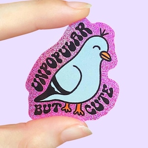 Könnte beinhalten: Ein glitzernder rosa Aufkleber mit einer hellblauen Taubenillustration und einem orangefarbenen Schnabel. Der schwarze Text um den Vogel herum lautet UNPOPULAR BUT CUTE in einer fetten, abgerundeten Schrift. Der Aufkleber hat eine unregelmäßige Form.