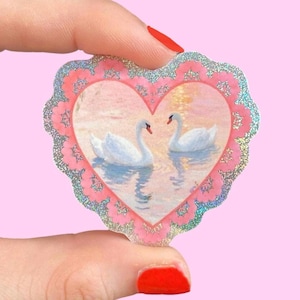 Könnte beinhalten: Ein herzförmiger Aufkleber mit einem Gemälde von zwei weißen Schwänen auf einem See. Der Aufkleber hat einen rosa Rand mit einem gewellten Rand und einen glitzernden silbernen Besatz. Der Hintergrund ist ein zartes Rosa.