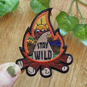 Könnte beinhalten: Ein gestickter Aufnäher in Form eines Lagerfeuers mit orangefarbenen und roten Flammen, braunen Holzscheiten und einem beigen Dreieck in der Mitte, das den schwarzen Text STAY WILD enthält. Das Design zeigt Sonne, Bäume und Berge.