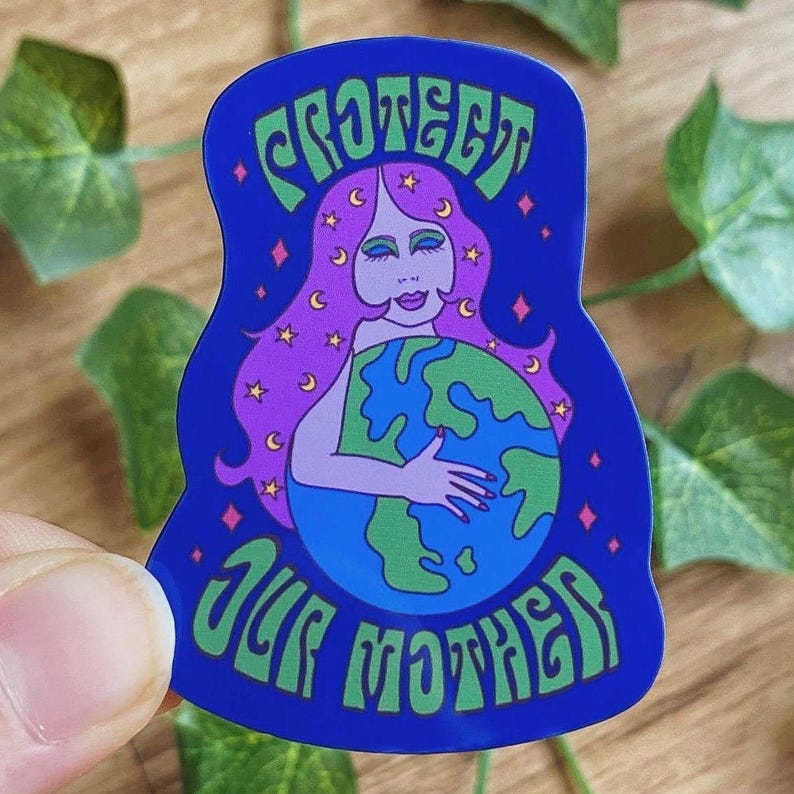 May include: A blue sticker with the words "PROTECT OUR MOTHER" in green. The sticker features a woman with pink hair, holding the Earth. The woman is surrounded by stars and moons. The sticker is held by a hand.