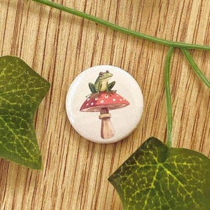 Könnte beinhalten: Ein runder Knopf mit einem grünen Frosch, der auf einem roten Pilz mit weißen Punkten sitzt. Der Knopf hat einen weißen Hintergrund und einen hellbraunen Stiel. Der Knopf ist ein skurriles Design.
