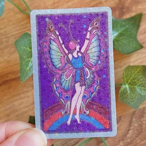 May include: A rectangular sticker with a purple background and a holographic effect. The sticker features a woman with butterfly wings, a blue dress, and a rainbow at the bottom. The woman is raising her arms, and the wings are pink and blue.