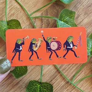 Puede incluir: Una pegatina rectangular naranja con una banda de marcha de cuatro ranas. Cada rana lleva un esmoquin negro y toca un instrumento diferente: trompeta, saxofón, tambor y batuta. Las ranas son verdes con vientres blancos.