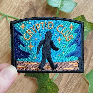以下が含まれることがあります： オレンジ色の「CRYPTID CLUB」の文字が入った刺繍パッチ。歩いている人の黒いシルエットが特徴です。背景はターコイズブルーで、オレンジ色の星と青い木が描かれています。パッチには黒い縁取りがあります。