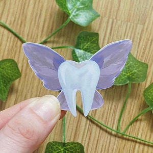 May include: A whimsical sticker featuring a tooth with lavender fairy wings. The tooth is white and light blue, with a detailed design. The wings have a gradient effect. The sticker is held by a hand, with green leaves in the background.