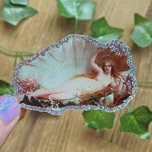 May include: A sticker depicting a classical painting of a nude woman in a seashell. The artwork uses a light colour palette with red coral accents and a glittery border. The woman has long red hair and wears a golden crown.