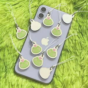 May include: A collection of frog-shaped phone charms with white lanyards. Each charm features a green frog face with a pink heart. Some charms are on a silver phone, others are separate. The background is a bright green, fuzzy surface.