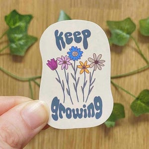 Könnte beinhalten: Ein cremefarbener Aufkleber mit den Worten "Keep Growing" in blauer Schrift, über einer farbenfrohen Illustration von Blumen. Die Blumen umfassen eine lila Tulpe, rosa, blaue, gelbe und beige gänseblümchenartige Blüten.