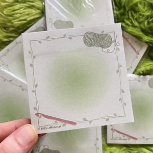 May include: Square notepad with a light green gradient background, a border of leaves, and illustrations of yarn and a crochet hook. The text "(c) 2025 LOUISEKIND" is printed at the bottom. The notepad is held in a hand.