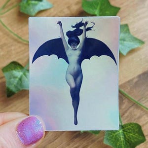 May include: A rectangular sticker featuring a nude figure with bat wings, arms raised. The figure is in shades of blue and gray, with dark hair. The background has a subtle iridescent effect. The sticker is held by a hand with a purple nail.