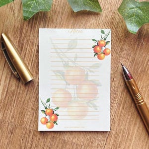 Könnte beinhalten: Notizblock mit Aquarell-Illustration von Orangen und Blättern, mit dem Wort "Notes" oben. Der linierte Notizblock liegt auf einer Holzoberfläche, zusammen mit zwei goldenen Stiften.