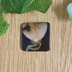 Könnte beinhalten: Quadratischer Knopf mit einem künstlerischen Bild eines Gesichts und einer Hand, die einen leuchtenden Gegenstand hält. Der Knopf hat einen schwarzen Rand und befindet sich auf einem Holzgrund mit grünen Blättern.