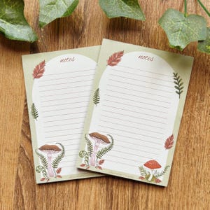 Può includere: Due fogli rettangolari per appunti con un bordo verde chiaro e un centro arcuato bianco contenente linee orizzontali. Il design presenta illustrazioni di funghi marroni, funghi rossi e foglie di felce verdi. La parola notes è stampata in alto.