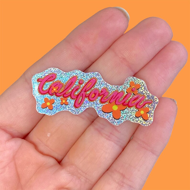 May include: A holographic sticker with the word "California" in pink script, accented by orange flowers. The sticker has a glittery, iridescent finish and is held in a hand against an orange background.