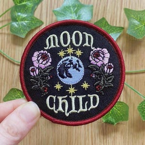 Könnte beinhalten: Runder, schwarz bestickter Aufnäher mit den cremefarbenen Worten "MOON CHILD". Der Aufnäher zeigt einen hellblauen Globus, rosa Rosen und goldene Sterne mit einem roten Rand. Grüne Blätter umgeben den Aufnäher.