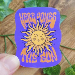 Könnte beinhalten: Ein violetter Aufkleber mit dem Text "HERE COMES THE SUN" in Orange, der eine Illustration einer lächelnden Sonne zeigt. Die Sonne hat gelbe Strahlen und ein Gesicht mit geschlossenen Augen. Der Aufkleber wird von einer Hand gehalten.