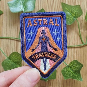 Könnte beinhalten: Gestickter Aufnäher mit blauem Hintergrund und den Worten "ASTRAL TRAVELER". Der Aufnäher zeigt eine Silhouette mit ausgestreckten Armen, umgeben von orangefarbenen und rosa Akzenten sowie vier weißen Kreuzen.