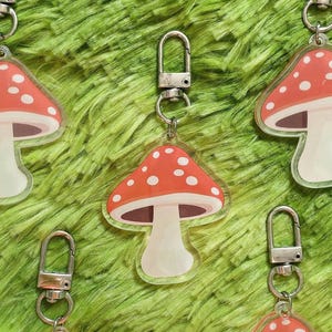 Könnte beinhalten: Schlüsselanhänger mit einem roten Pilzdesign mit weißen Punkten. Der Pilz hat einen weißen Stiel und ist an einem silberfarbenen Metallverschluss befestigt. Die Schlüsselanhänger befinden sich auf einem strukturierten grünen Hintergrund.