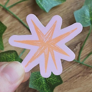 Peut inclure: Un autocollant avec un motif d'éclat d'étoile orange sur un fond rose clair. L'autocollant est tenu par les doigts d'une personne. Le motif d'éclat d'étoile a un aspect dessiné à la main.