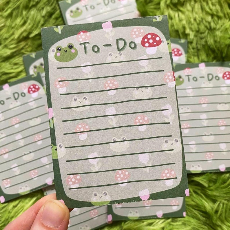 May include: A stack of To-Do list notepads with a green border and a light green background. The notepads feature a repeating pattern of cartoon frogs and mushrooms. The words "To-Do" are printed at the top of each notepad.