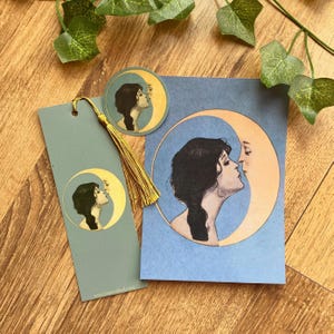 Könnte beinhalten: Ein Schreibwarenset mit dem Profil einer Frau, die einen Halbmond küsst. Das Set enthält einen Lesezeichen mit einer goldenen Quaste, einen Aufkleber und eine Karte. Das Kunstwerk ist in Blau-, Gold- und Schwarztönen gehalten.