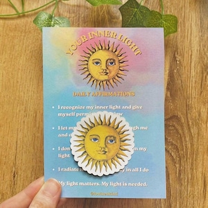 Könnte beinhalten: Eine rechteckige Karte mit der Aufschrift Your Inner Light Daily Affirmations und einer gelben Sonnenillustration mit Gesicht. Ein runder Aufkleber mit demselben Sonnendesign ist über dem Text platziert. Die Karte hat einen blau-violetten Verlauf.