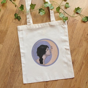 Könnte beinhalten: Cremefarbene Tragetasche mit einer kreisförmigen Illustration eines Frauenprofils, das ein anderes Profil küsst. Die Illustration ist vor einem hellvioletten und pfirsichfarbenen Hintergrund platziert. Die Tasche hat zwei lange Griffe.