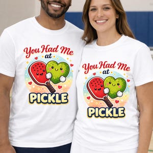 Peut inclure: T-shirts blancs avec un graphisme coloré. Le graphisme représente une raquette de pickleball rouge et un cœur vert qui s'embrassent, avec le texte "You Had Me at PICKLE". Le motif comprend des cœurs et un fond arc-en-ciel.