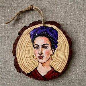 Puede incluir: Un adorno redondo de madera con un retrato pintado de Frida Kahlo. Lleva una diadema morada y tiene el pelo oscuro. El fondo es un patrón de veta de madera.