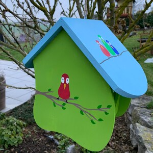 May include: A wooden birdhouse with a light blue roof and a lime green body. The front features a painted red bird perched on a brown branch with green leaves. The roof displays three small painted birds in red, green, and blue on a branch.