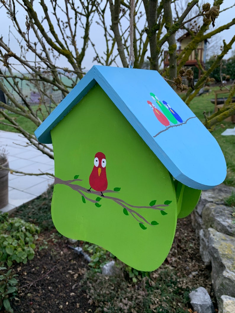 May include: A wooden birdhouse with a light blue roof and a lime green body. The front features a painted red bird perched on a brown branch with green leaves. The roof displays three small painted birds in red, green, and blue on a branch.