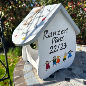 May include: A white wooden house-shaped bird feeder featuring the black text "Ranzen P&auml;nz 22/23" on the front, accompanied by a colorful illustration of five children. The roof displays various handwritten names, a small bee, and ladybug illustrations.