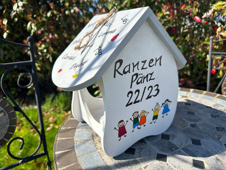 May include: A white wooden house-shaped bird feeder featuring the black text "Ranzen P&auml;nz 22/23" on the front, accompanied by a colorful illustration of five children. The roof displays various handwritten names, a small bee, and ladybug illustrations.