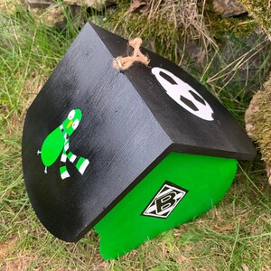 May include: A black and green wooden birdhouse featuring a painted green bird wearing a black and white striped scarf on the side. The roof is black with a white circular graphic, and a diamond-shaped logo with the letter B is on the green wall.