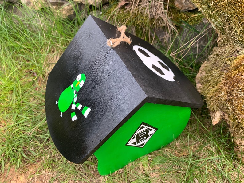 May include: A black and green wooden birdhouse featuring a painted green bird wearing a black and white striped scarf on the side. The roof is black with a white circular graphic, and a diamond-shaped logo with the letter B is on the green wall.
