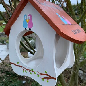 May include: A white wooden birdhouse with a brown roof, featuring hand-painted illustrations of a blue and pink bird pair above the circular entrance. A brown branch with green leaves is painted on the front, and the date 23.08.2020 is visible.