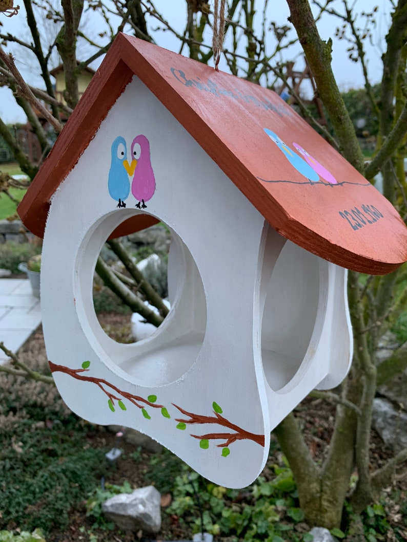 May include: A white wooden birdhouse with a brown roof, featuring hand-painted illustrations of a blue and pink bird pair above the circular entrance. A brown branch with green leaves is painted on the front, and the date 23.08.2020 is visible.