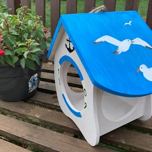 May include: A white wooden birdhouse with a bright blue roof featuring painted white seagulls. The front has a circular opening surrounded by a blue ring, a black anchor icon at the top, and the numbers 1, 8, 0, and 9 printed around the edge.