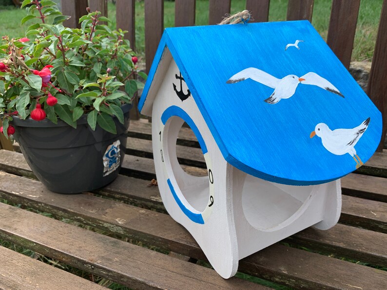 May include: A white wooden birdhouse with a bright blue roof featuring painted white seagulls. The front has a circular opening surrounded by a blue ring, a black anchor icon at the top, and the numbers 1, 8, 0, and 9 printed around the edge.